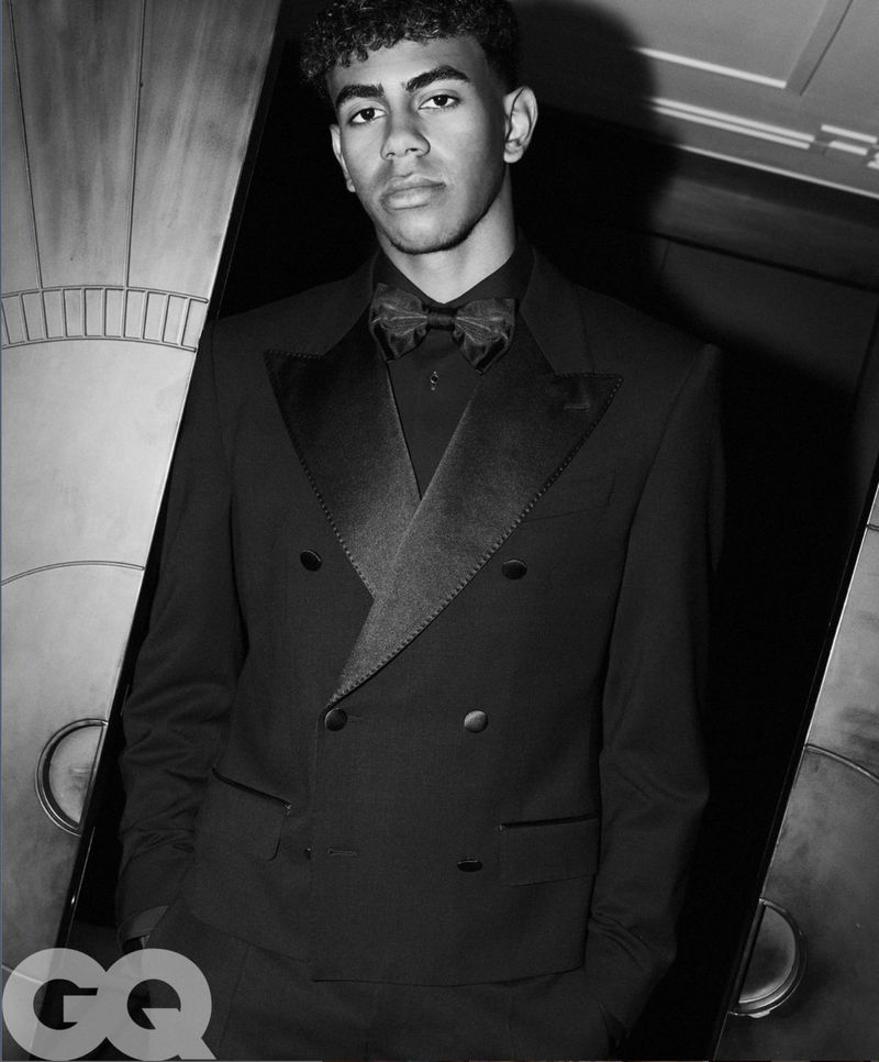 Monochrome portrait of a young adult male in a black tuxedo with a bow tie, looking at the camera, in a dark indoor setting.