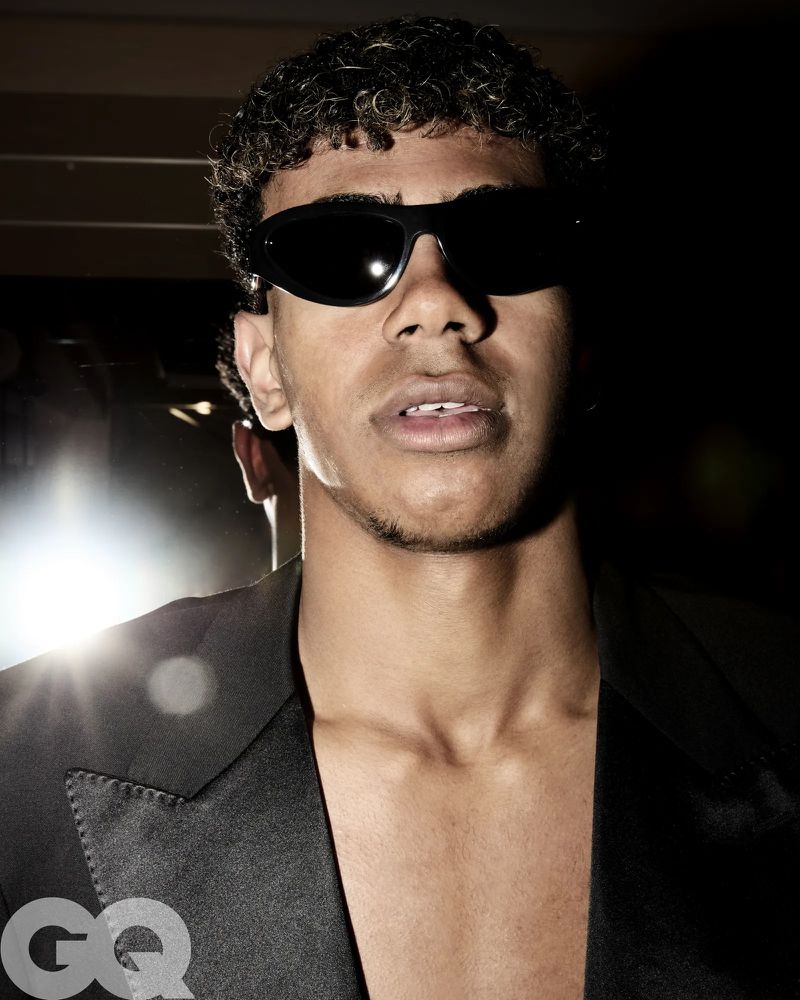 Close-up portrait of a stylish person wearing sunglasses and a black jacket against a dark background with studio lighting and GQ logo.
