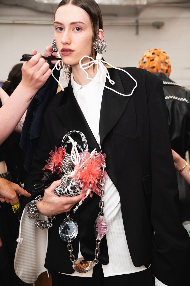A fair-skinned young adult woman stands indoors, wearing an oversized black blazer and decorative jewelry, while a hand applies makeup.
