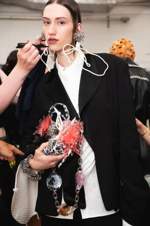 A fair-skinned young adult woman stands indoors, wearing an oversized black blazer and decorative jewelry, while a hand applies makeup.