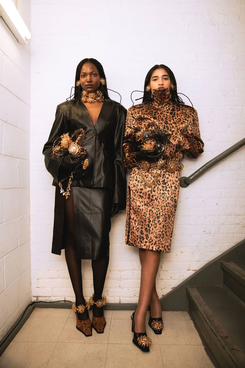 Two women pose in a stairwell wearing leopard and leather dresses with gold jewelry, purses, and shoes. High fashion photoshoot.
