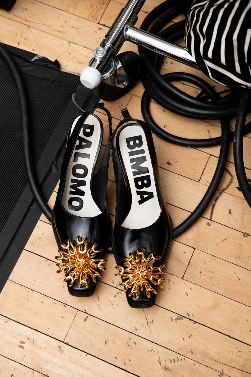 Close-up of black slingback shoes with gold sunburst embellishments against a backdrop of lighting equipment and wooden flooring.