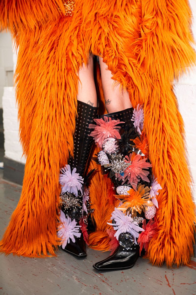 Legs in floral-embellished black boots are framed by a fuzzy orange coat, standing on a concrete floor against a plain white wall.