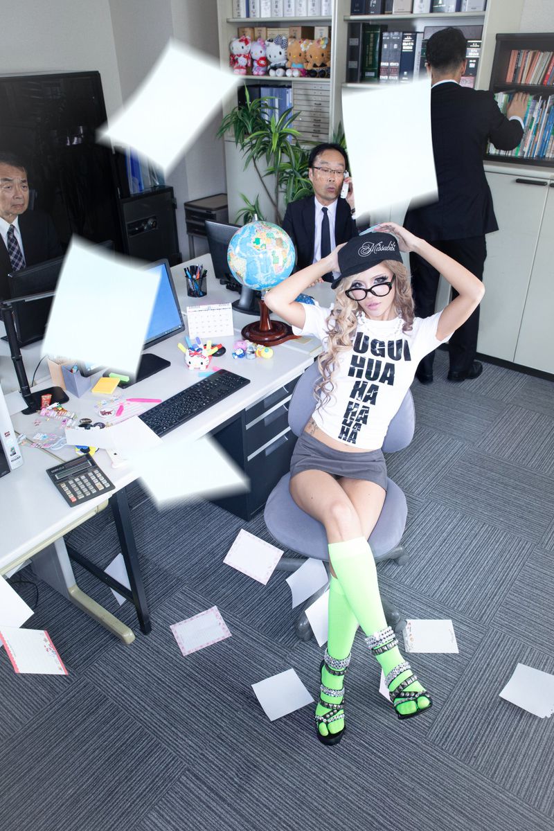 Young woman sits in an office chair surrounded by flying papers, colleagues working in the background, and a desk with office supplies.