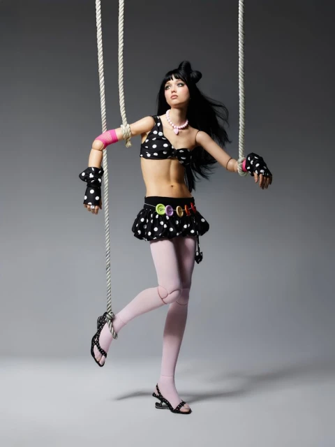 Young woman in polka dot outfit poses as a puppet against gray background. Strings attached to her limbs create a unique fashion statement.