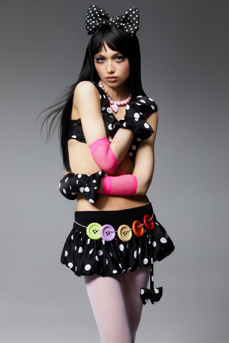 Young woman in a polka-dot outfit poses against a gray background. She is wearing a black wig with bangs and a large hair bow.