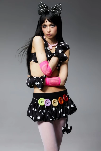 Young woman in a polka-dot outfit poses against a gray background. She is wearing a black wig with bangs and a large hair bow.