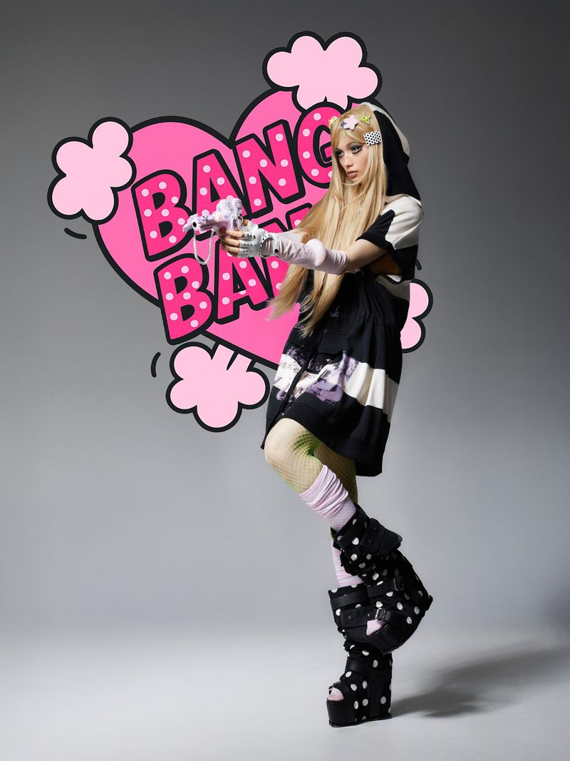 Blonde woman in a black and white outfit posing with a toy gun in front of a pink heart-shaped cartoon graphic on grey background.