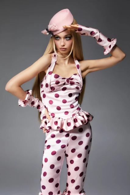 Young woman poses in light pink and burgundy polka dot top, pants, hat, and gloves against a grey backdrop, studio lighting.