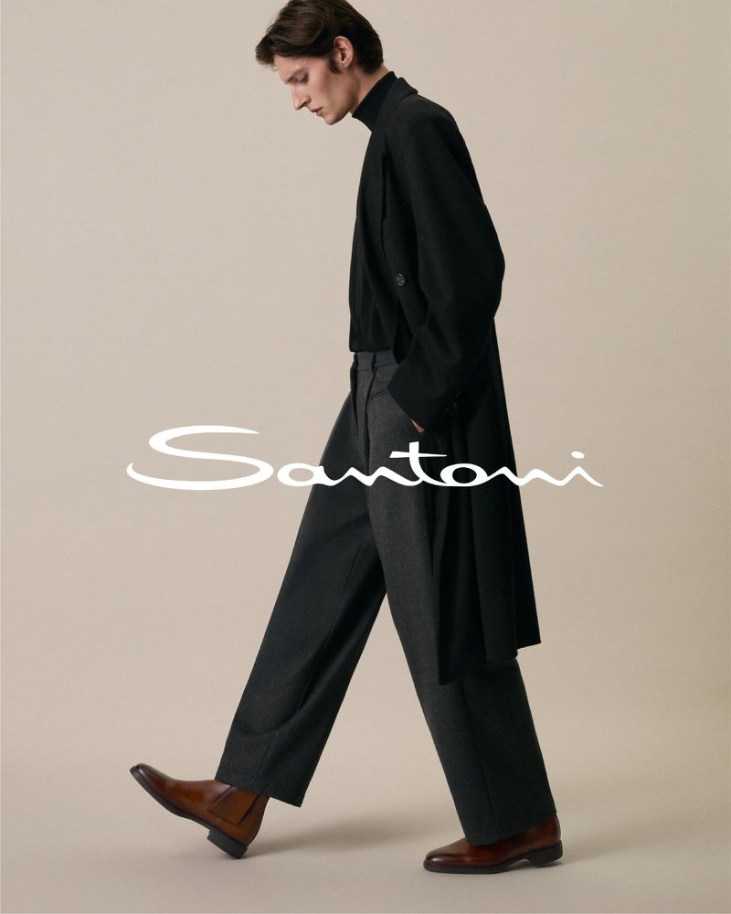 Editorial fashion shot of a slim adult man in a black coat and grey pants, walking and modeling brown leather dress shoes.