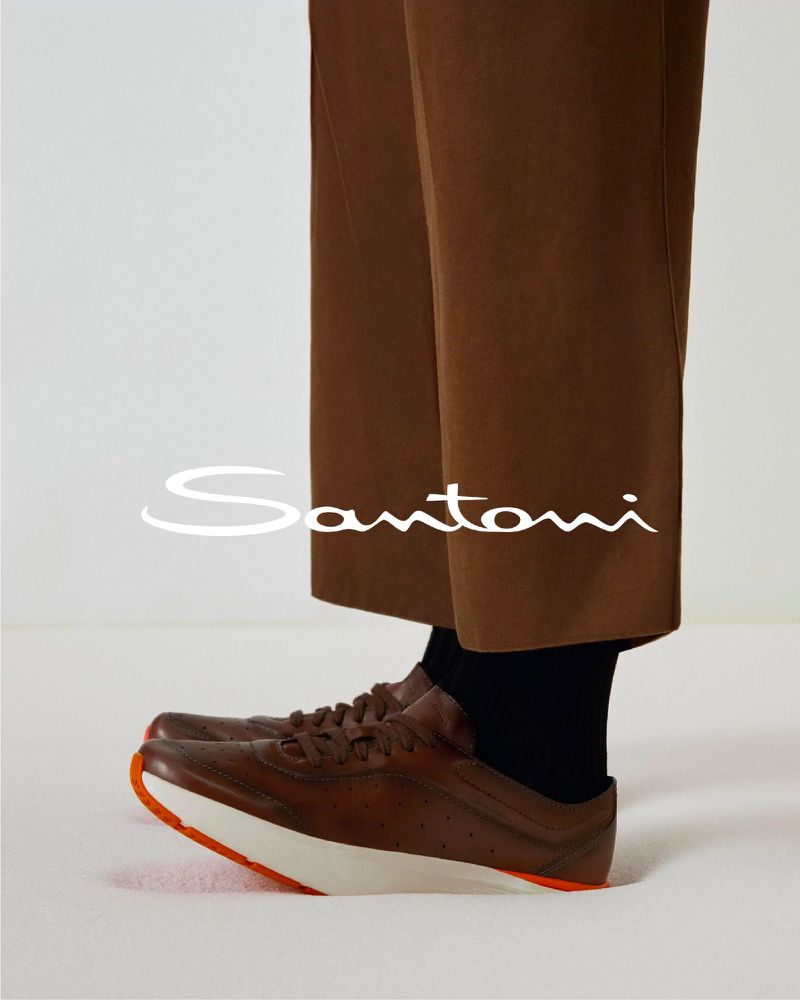 Close-up of Santoni brown leather sneakers with white soles, contrasted by brown dress pants and black socks on a white background.