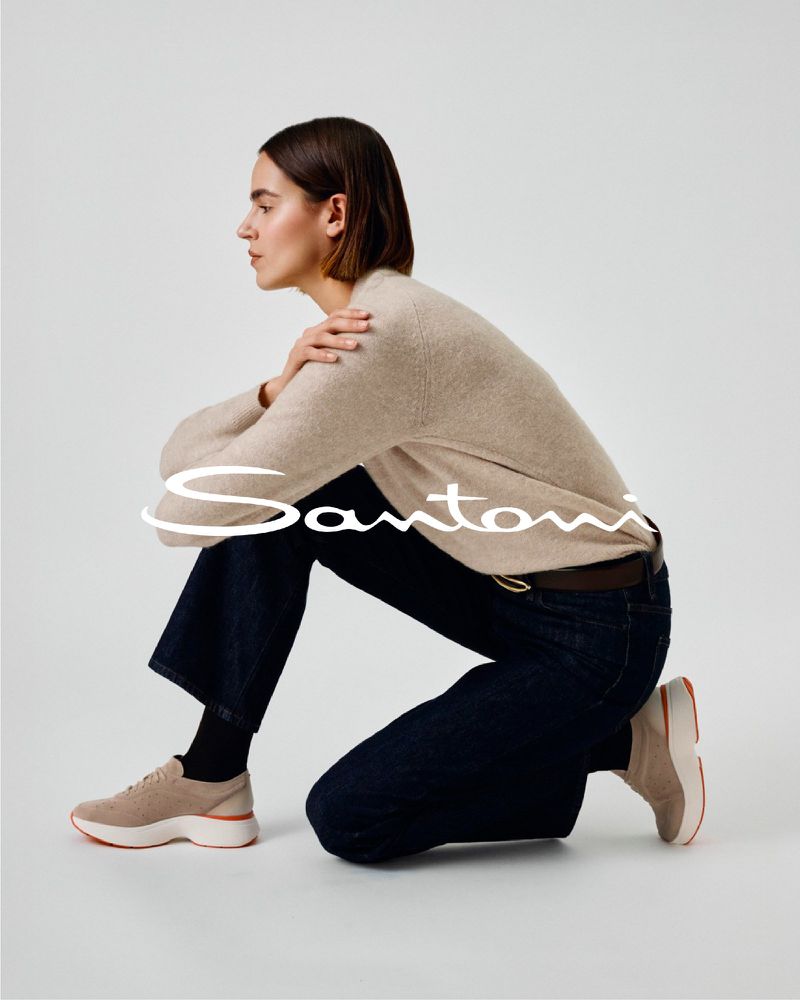Fashion editorial shot of a woman in a beige sweater and dark jeans crouching in a studio with Santoni logo overlay.