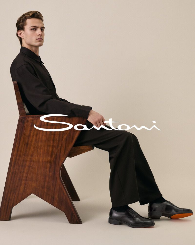 A man in an all-black outfit seated in a geometric wooden armchair wearing black leather dress shoes against a warm neutral studio backdrop.