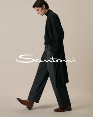 Model wearing a long black coat, charcoal wide-leg trousers, and brown leather boots walking against a beige studio backdrop.