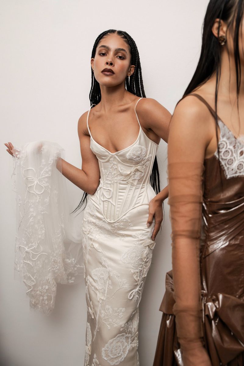 Woman in a corset-style white gown posing next to a partially visible woman in brown at a fashion shoot against a white backdrop.