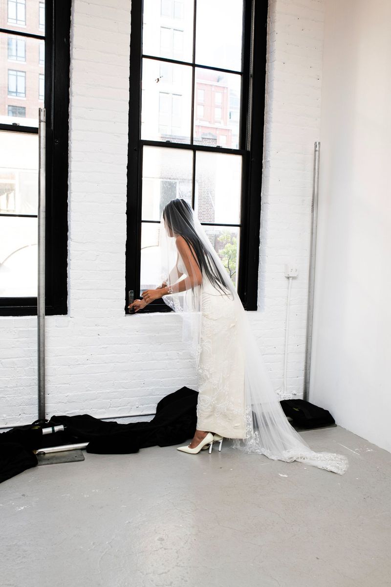 Elegant bride in white gown and veil stands by window in studio, exuding classic beauty and grace in bright, minimalist setting.