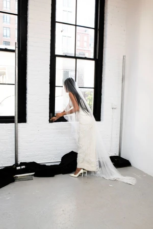 Elegant bride in white gown and veil stands by window in studio, exuding classic beauty and grace in bright, minimalist setting.