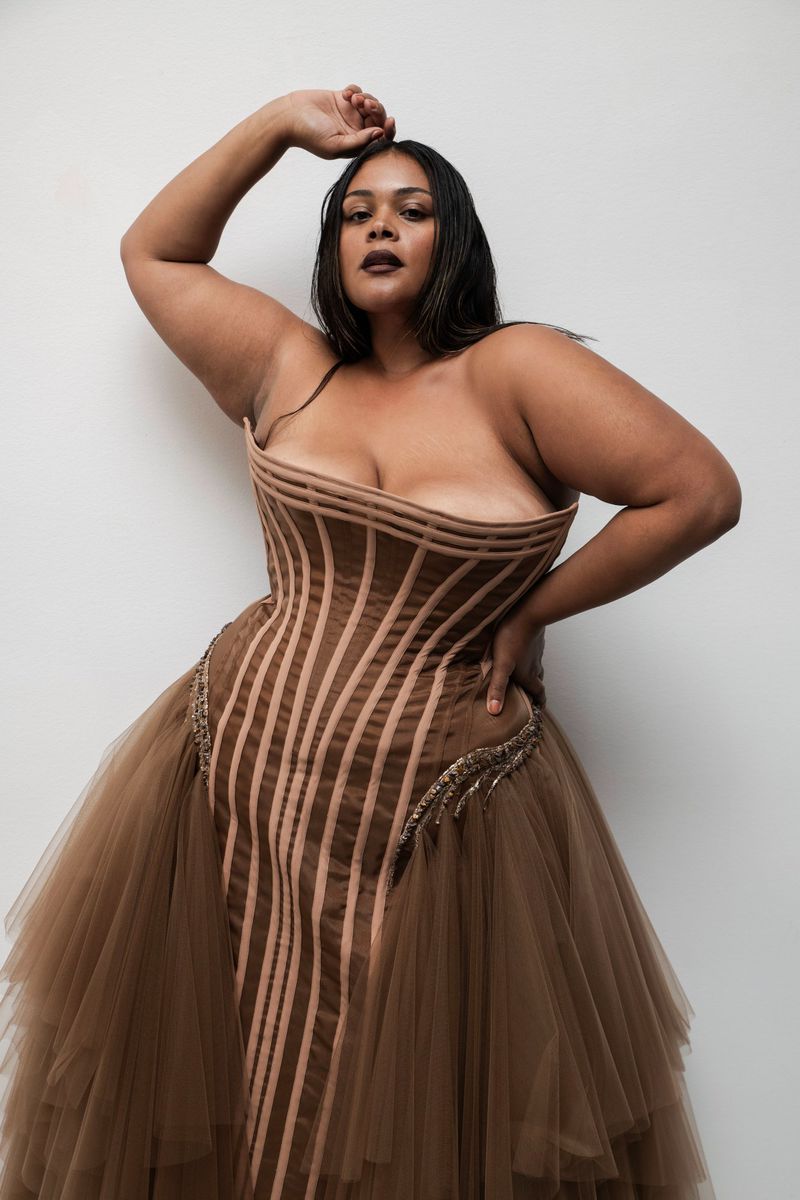 Adult woman in a strapless corset gown poses in a studio, with dark hair and neutral makeup.  Fashion editorial shot with a bright white background.