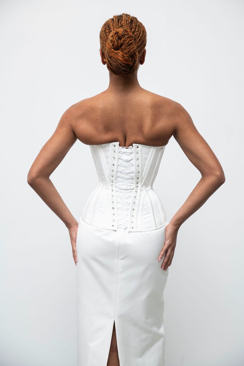 Rear view of a woman with auburn hair in a high bun, wearing a white corset and skirt against a bright white background.