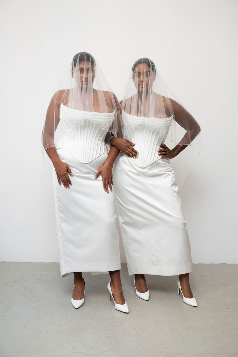 Two adult females in white dresses and veils pose indoors against a white wall, showcasing elegant minimalist fashion and formal wear.