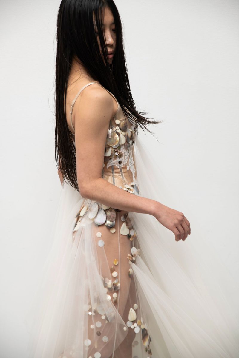 Asian woman in sheer gown adorned with seashells and beads, stands in a side profile against a stark white background. Fashion concept.