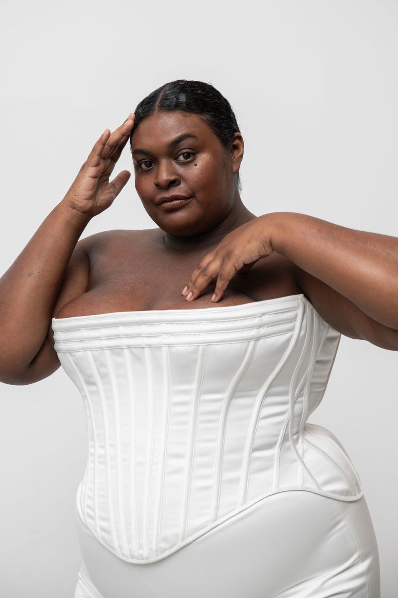 A dark-skinned woman in a white corset poses against a white background, fingers resting on her chest and head in a fashion shot.