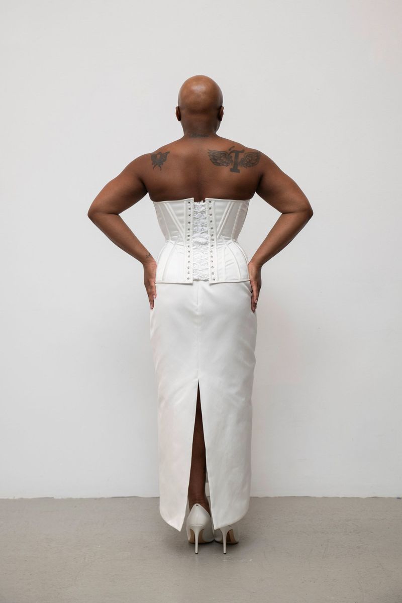 Model seen from behind wearing a structured white corset and matching skirt against a plain studio background.