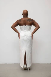 Model seen from behind wearing a structured white corset and matching skirt against a plain studio background.