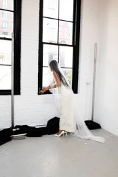 Model in a white bridal gown and long veil standing by a large multi-pane window in a studio setting