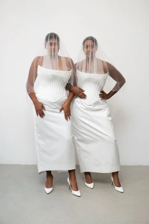 Two models in white structured corsets and long skirts wearing sheer veils standing against a plain backdrop.