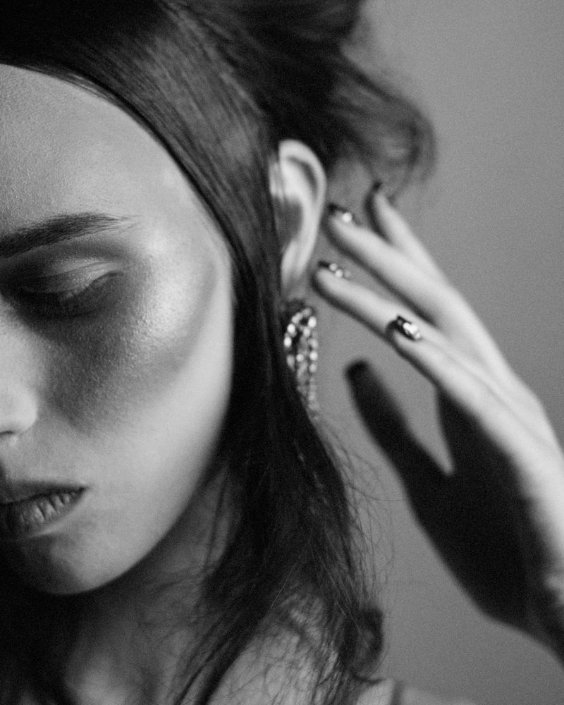 Monochrome portrait of an adult woman with dark hair and long earrings, posing with a hand near her face, against a solid gray background.
