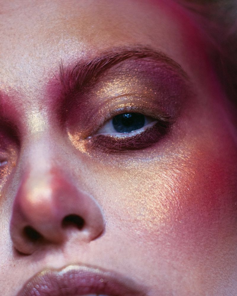 Close-up shot of a woman's face with bright pink and gold makeup emphasizing the eyes and skin, creating a vibrant beauty look.