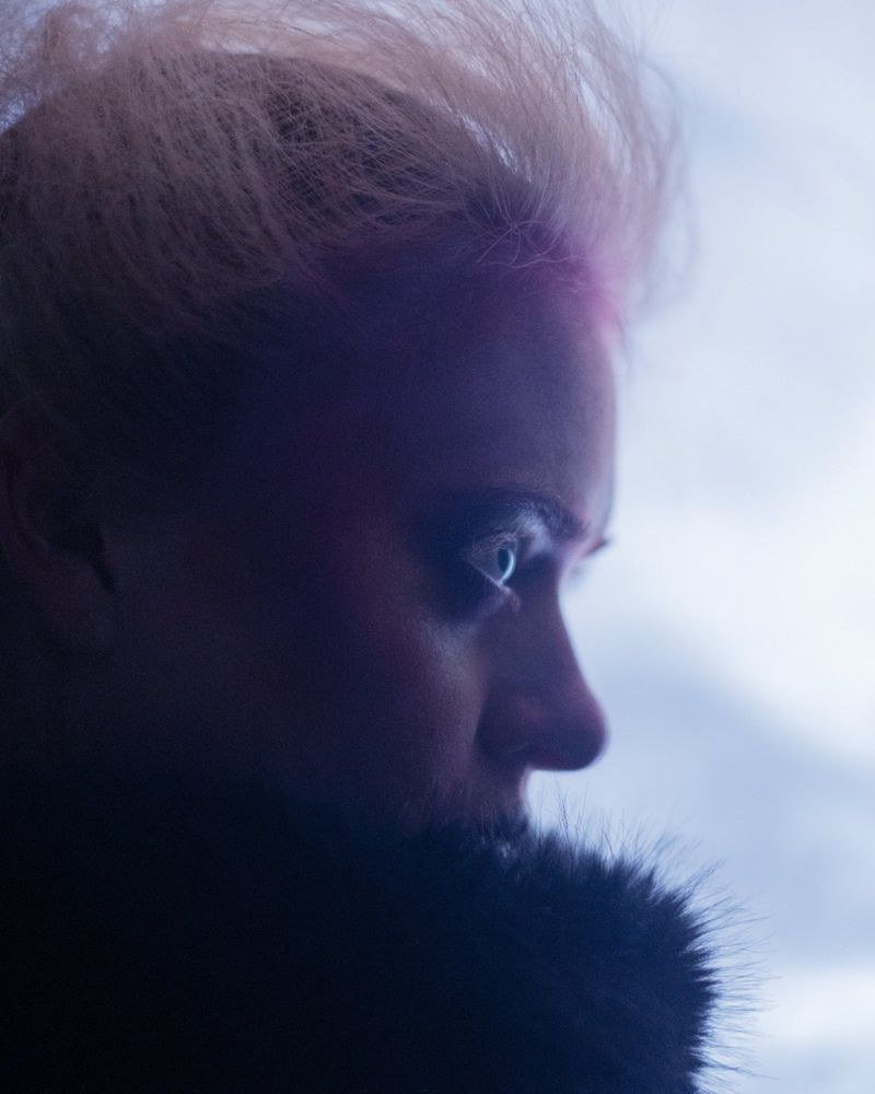 Close-up of a woman with styled blonde hair and light eyes looking off to the side, wearing a dark fuzzy collar in a neutral outdoor setting.