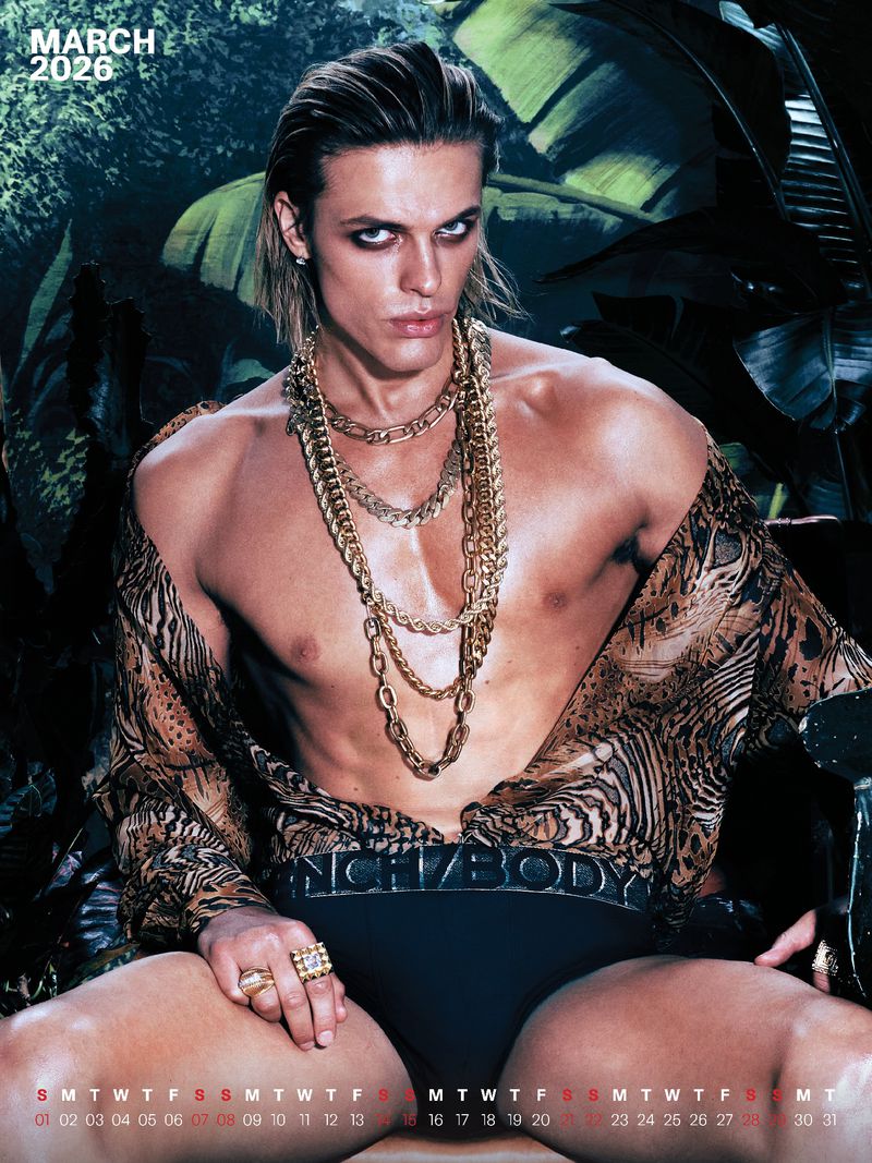 A male fashion model poses with a shirtless upper body and multiple gold chain necklaces in a jungle-themed setting for a calendar image.