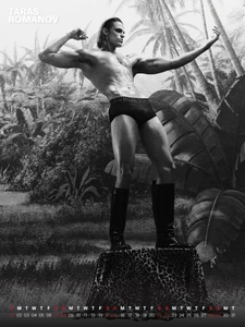 Model Taras Romanov in black briefs and tall boots posing on a leopard print pedestal in front of a painted jungle backdrop.