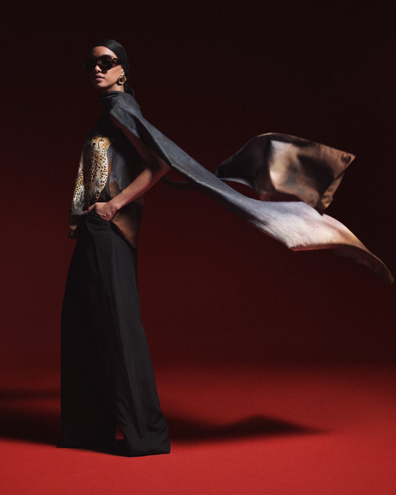 Model wearing black wide-leg trousers, patterned top, and a long flowing cape against a deep red studio backdrop.