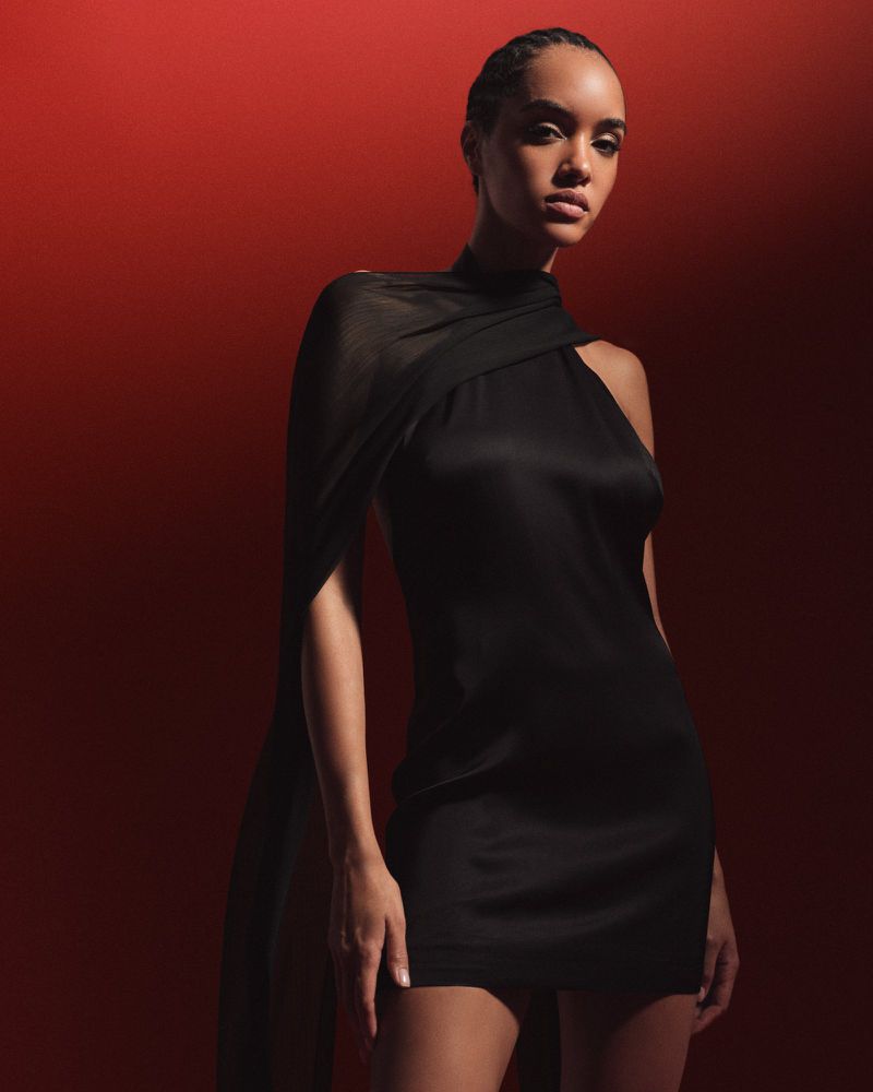 Model wearing a black mini dress with an asymmetric draped cape against a deep red background