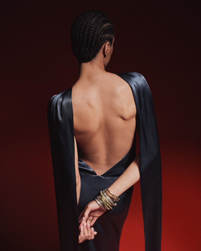 Back view of a woman with braided hair wearing a black open-back dress and bracelets in a studio with a red background. Fashion.
