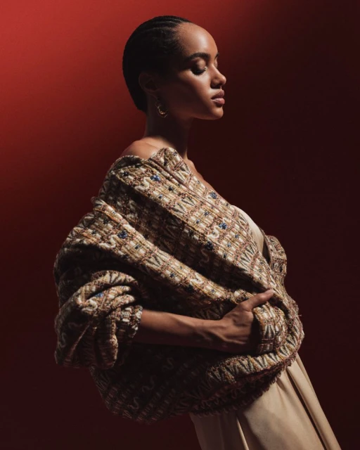 Elegant woman in off-shoulder knit shawl with intricate pattern, posed against a muted red backdrop in a fashion forward portrait.