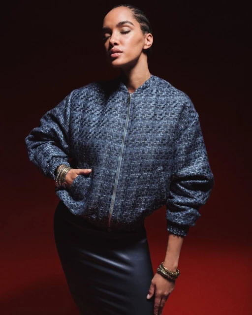 Woman posing in a bomber jacket and skirt against a red background. The jacket is blue with a textured pattern and shiny zipper.