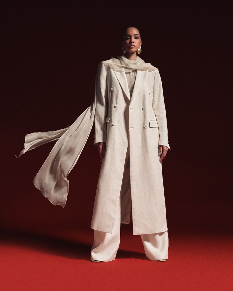 Model wearing a long cream tailored coat and wide-leg trousers with a flowing scarf against a red backdrop.