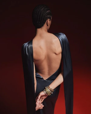 Rear view of a model wearing a backless dark satin gown with long draped sleeves and stacked bangles against a dark red backdrop.