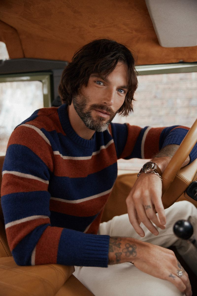 Man in striped sweater sits inside a car with tan interior, wearing a watch and rings, with tattoos on his arms, looking at the camera.