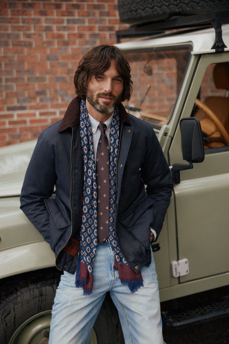 Man in navy jacket and jeans standing beside a Land Rover in front of a brick wall, wearing patterned scarf and tie, and brown hair.