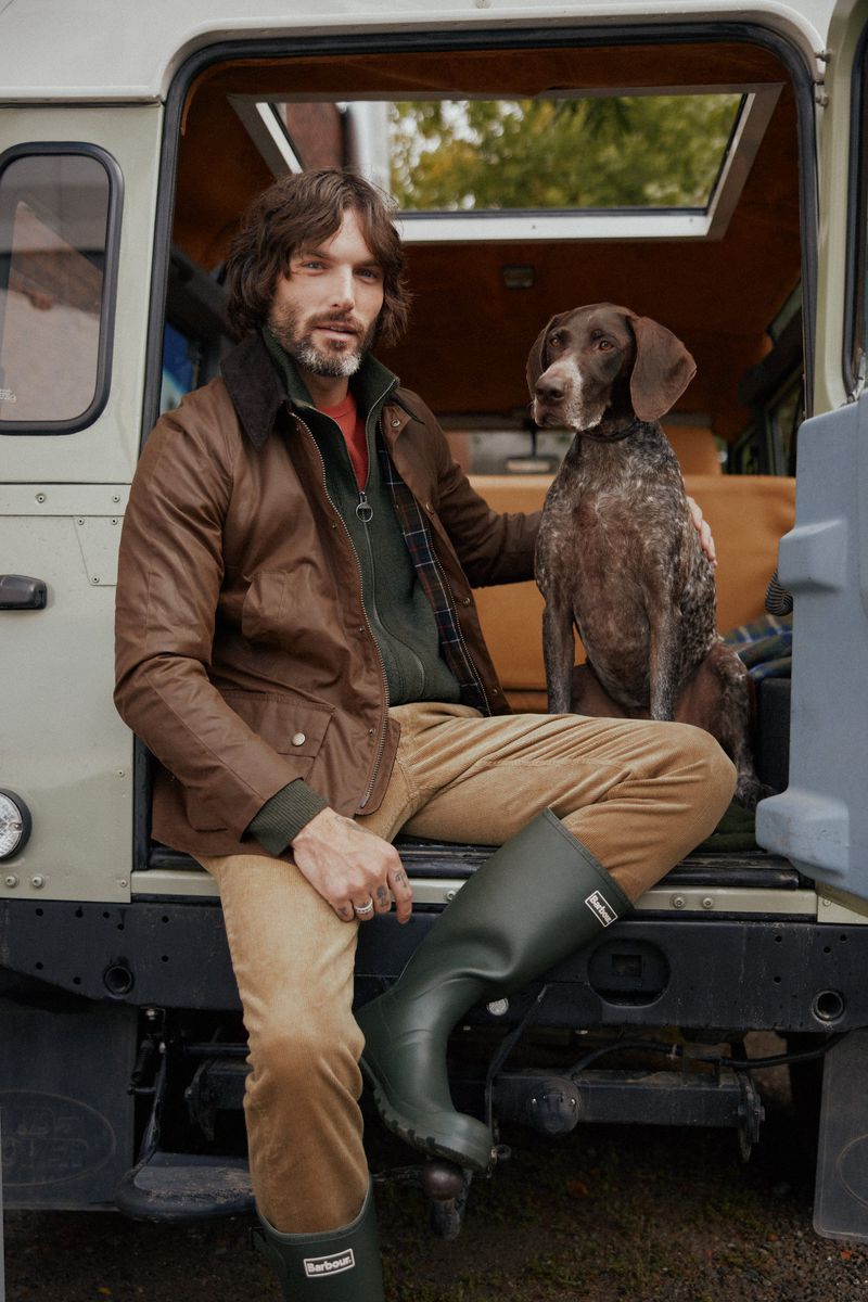 Man and dog sit on the back of a car. The man wears a wax jacket and tall rubber boots. Outdoor daytime shot. Barbour logo on the boots.