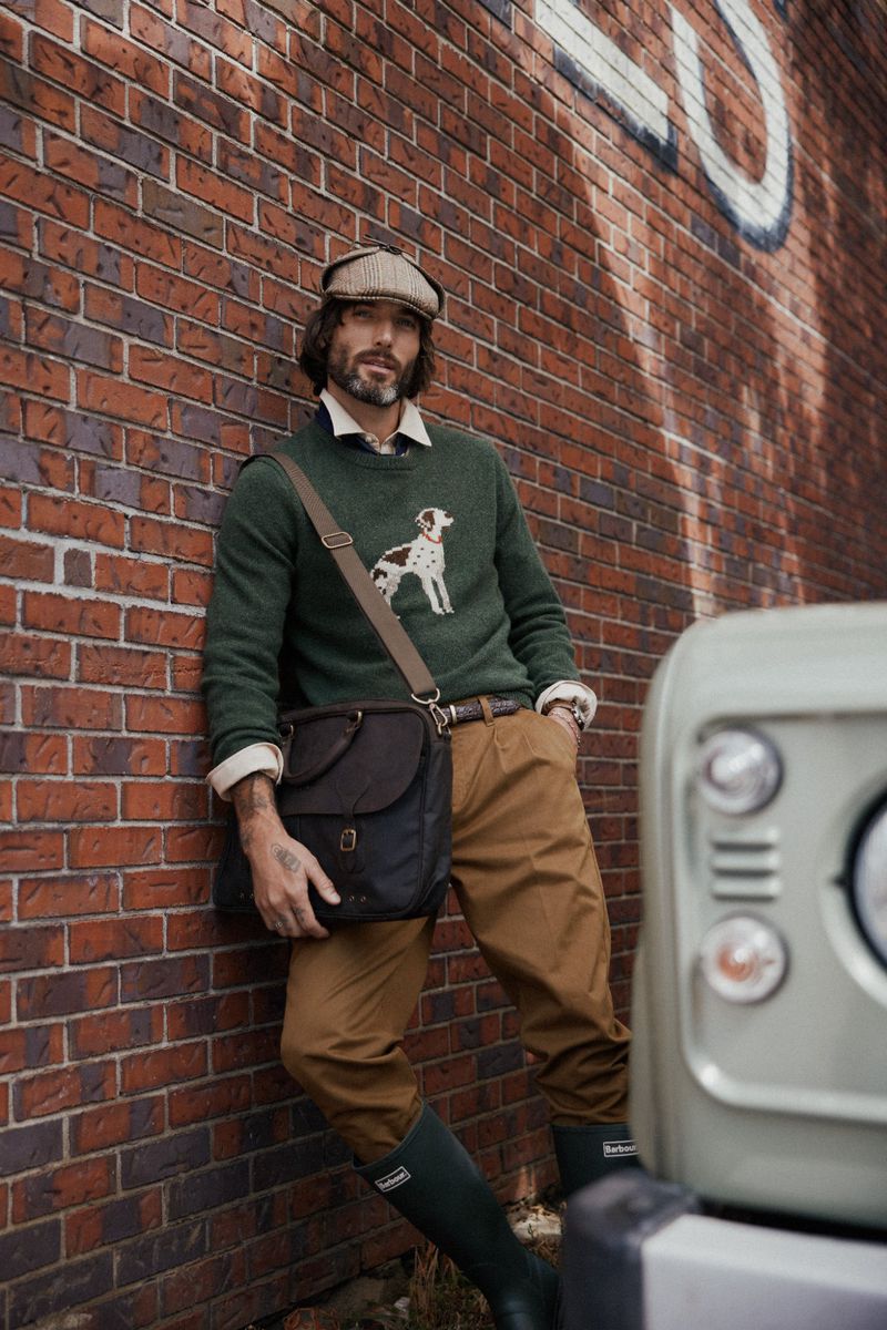 Stylish man with beard poses against brick wall next to vehicle wearing sweater with dog graphic and carrying shoulder bag.