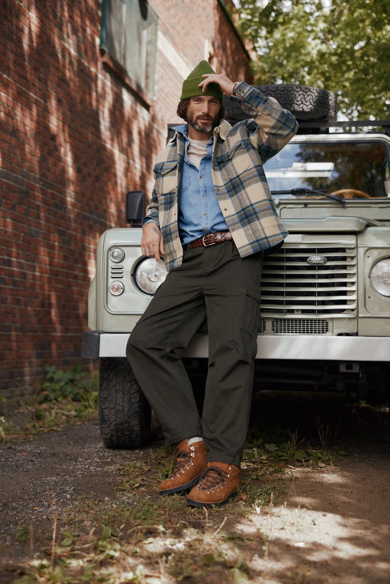A man poses leaning on a vintage 4x4 truck wearing a plaid shirt jacket, pants, beanie, and boots on a gravel path.