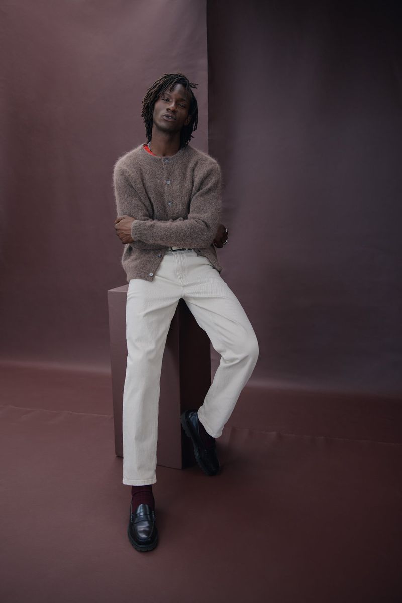 Adult man with dreadlocks wearing a brown cardigan, white pants, and black loafers posing in front of a brown background.
