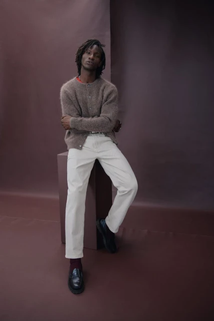 Adult man with dreadlocks wearing a brown cardigan, white pants, and black loafers posing in front of a brown background.