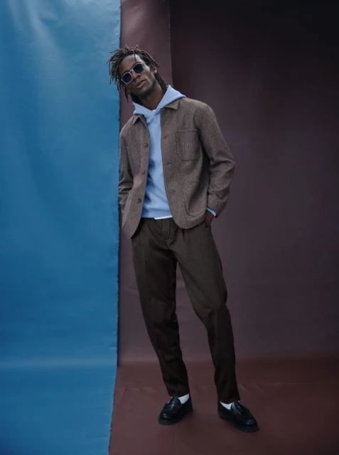 A man poses in a light-blue hoodie, brown jacket, and dark pants against a dual-tone backdrop in a studio shot.